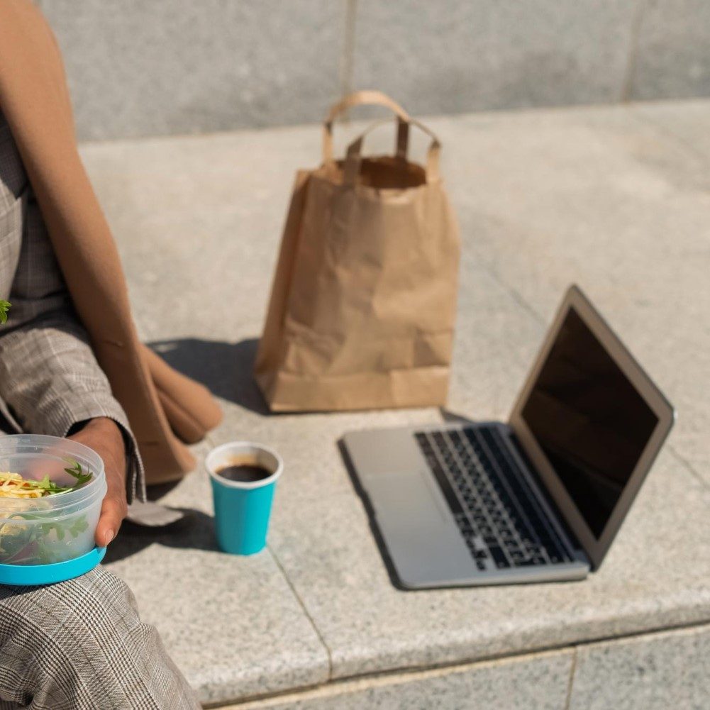 business person eating lunch next to laptop and brown lunch bag